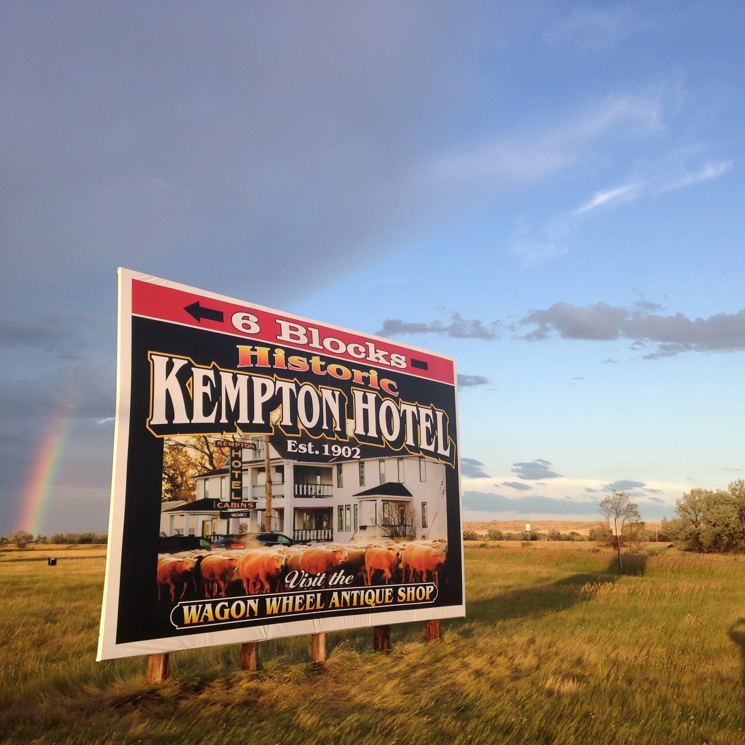A roadside billboard on the prairie pointing six blocks to the Historic Kempton Hotel, with a rainbow behind it