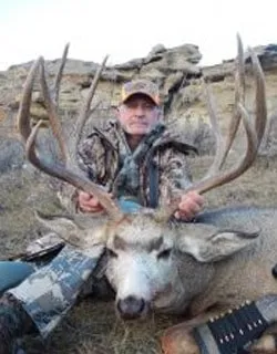 A hunter in camouflage kneeling behind a large mule deer buck on a rocky prairie hillside