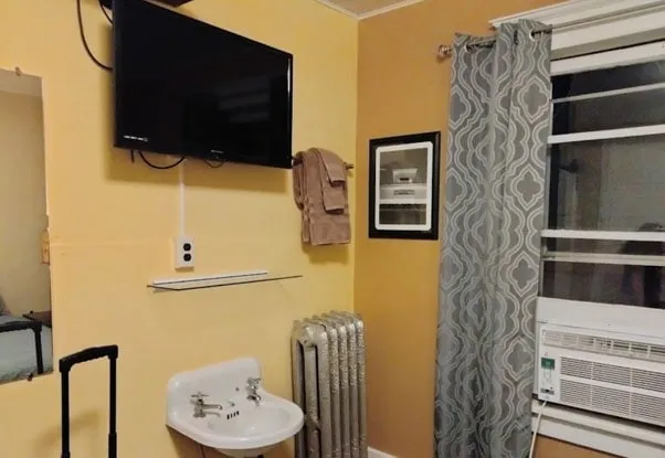 A historic economy room with a small corner sink, cast-iron radiator, window air conditioner and wall-mounted TV